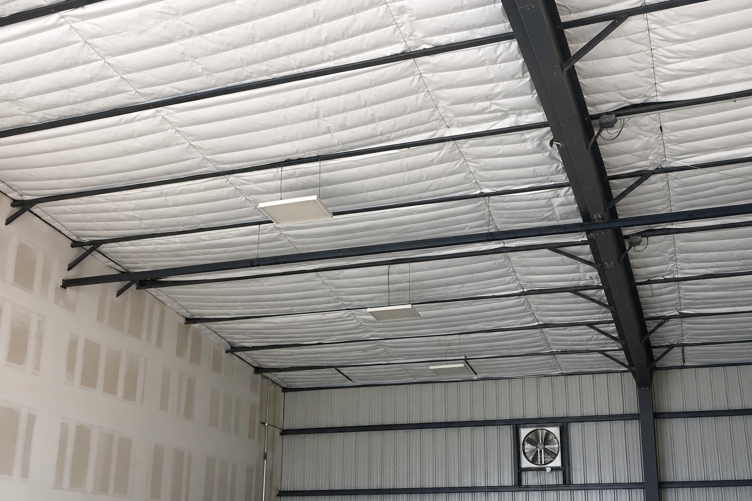 Commercial insulation lines a warehouse ceiling in white panels between dark steel beams.