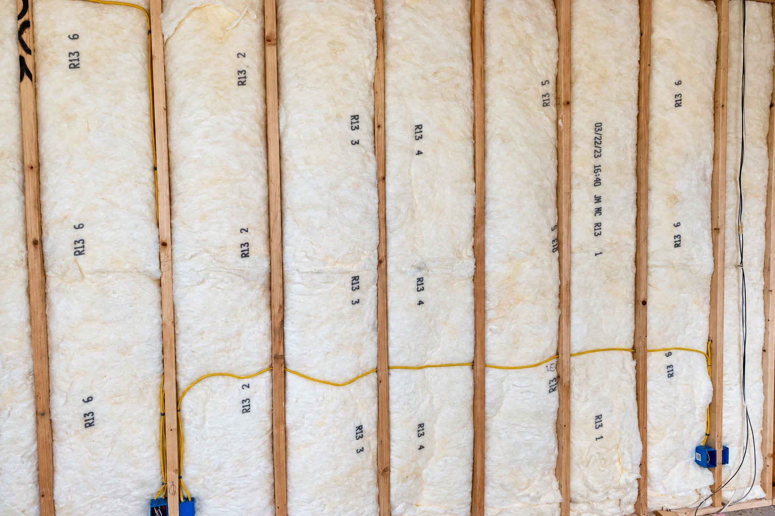 White fiberglass insulation fills wall cavities between exposed wooden studs, with electrical wiring visible.
