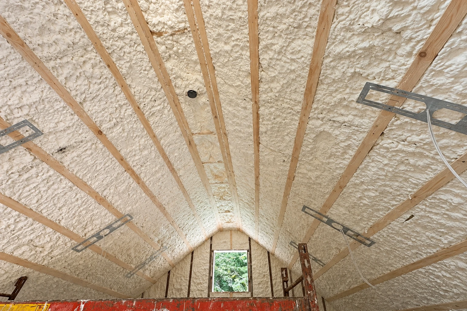 Spray foam insulation coats the sloped roof between exposed wooden rafters in an unfinished attic.