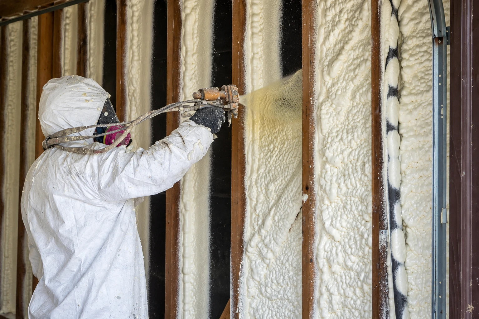 A worker in protective gear sprays foam insulation into wall cavities between exposed wooden studs.